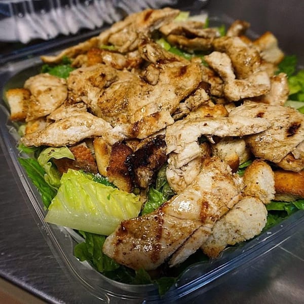 Grilled chicken and croutons on a bed of fresh green lettuce in a clear plastic container.