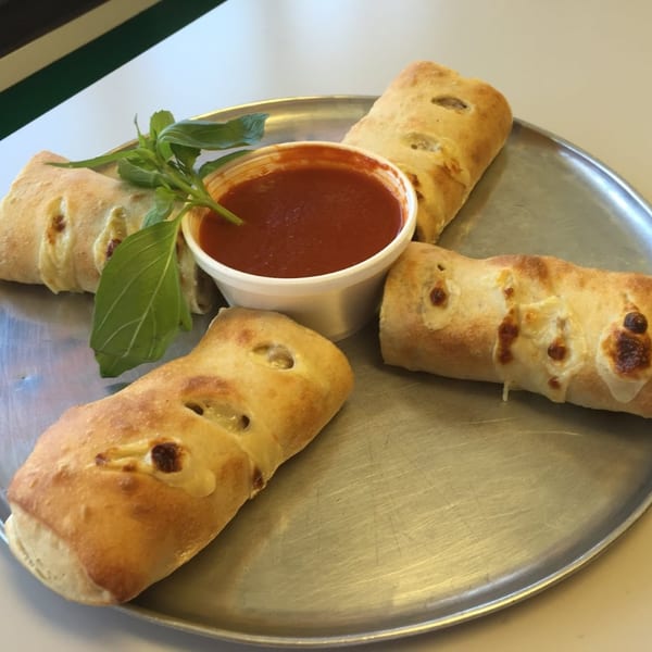 Four golden-brown calzone rolls with melted cheese, basil, and a cup of marinara sauce on a metal tray.