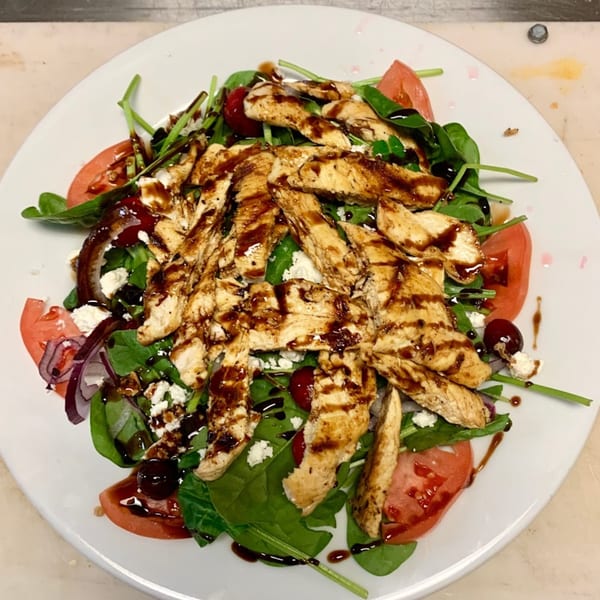 Grilled chicken salad with spinach, tomatoes, red onion, feta, and balsamic glaze on a white plate.